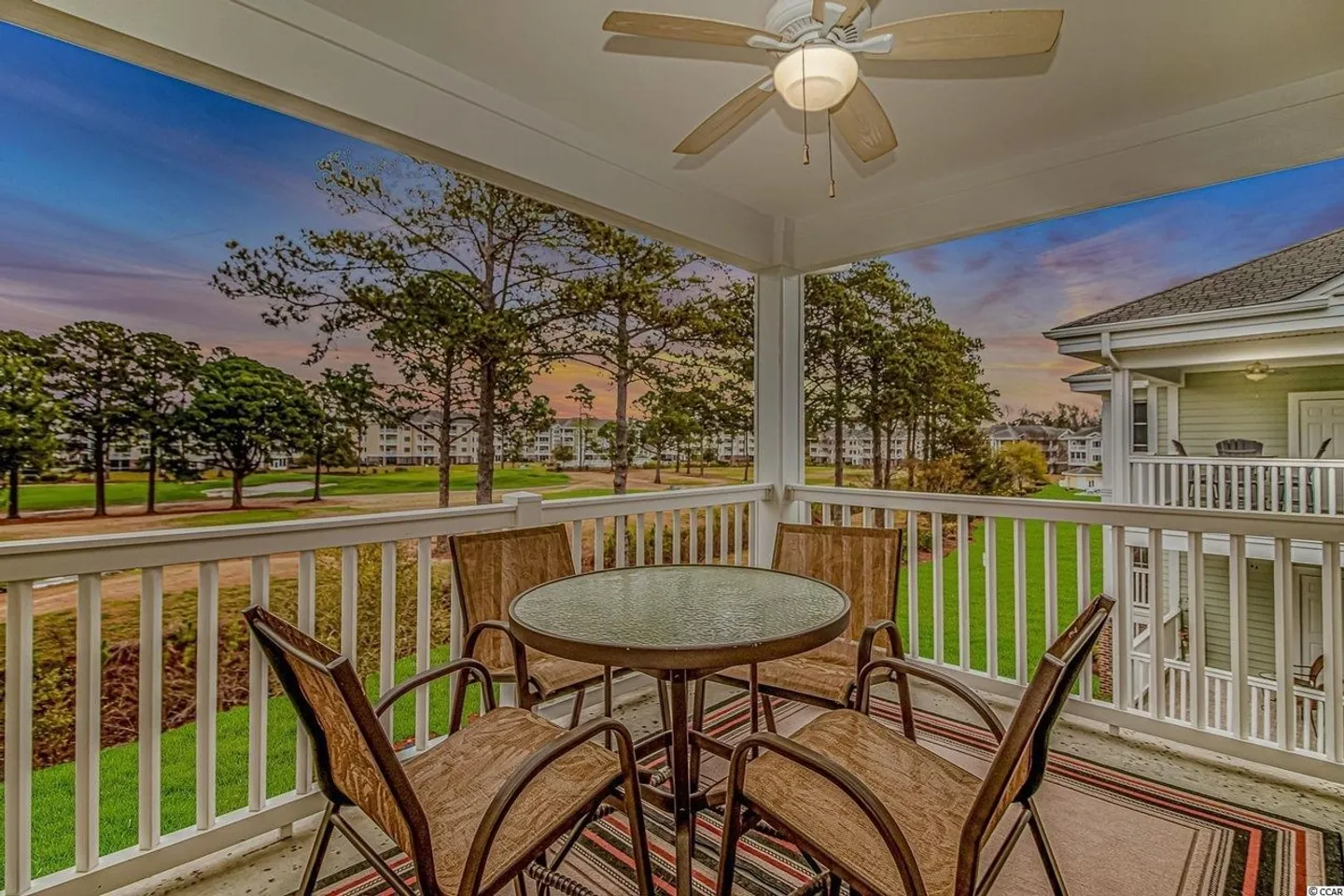 A covered balcony with a round table and four chairs overlooking trees and a green golf course area at sunset.