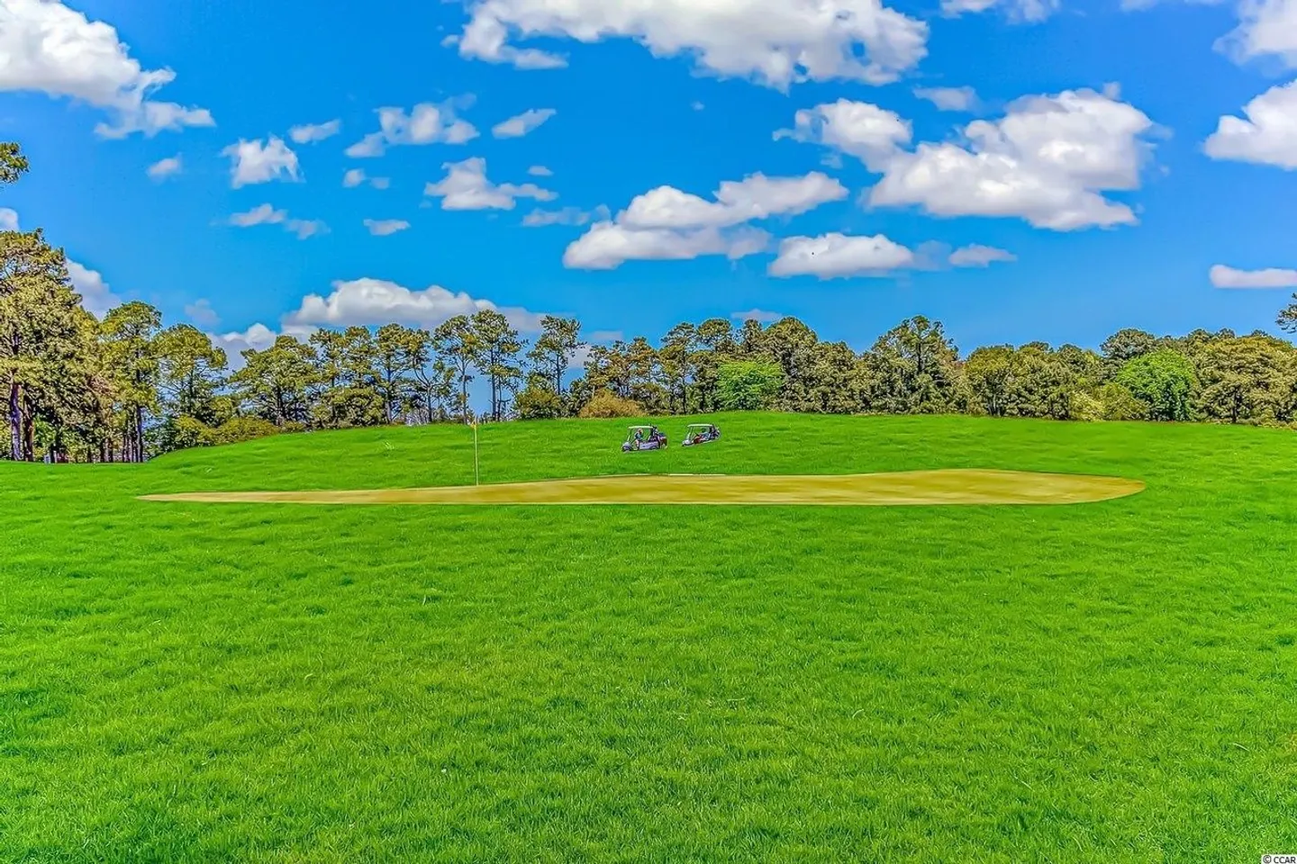 A golf course putting green and fairway under a bright blue sky with scattered clouds.
