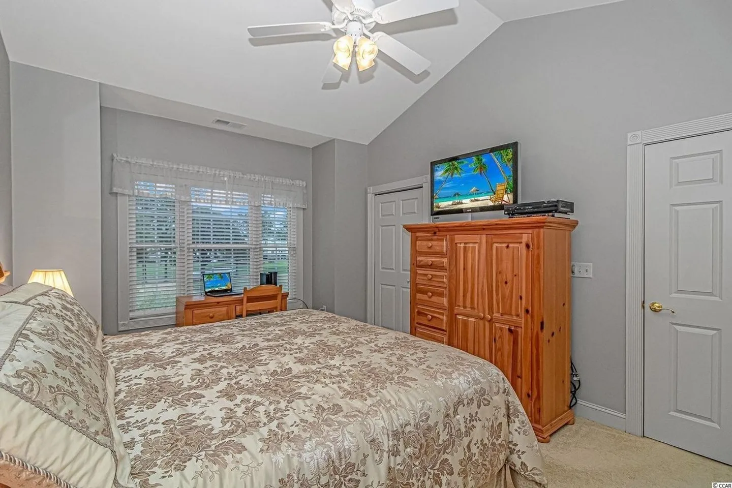 Bedroom with vaulted ceiling, ceiling fan, bed with patterned comforter, wooden armoire with television, and small desk by the window.