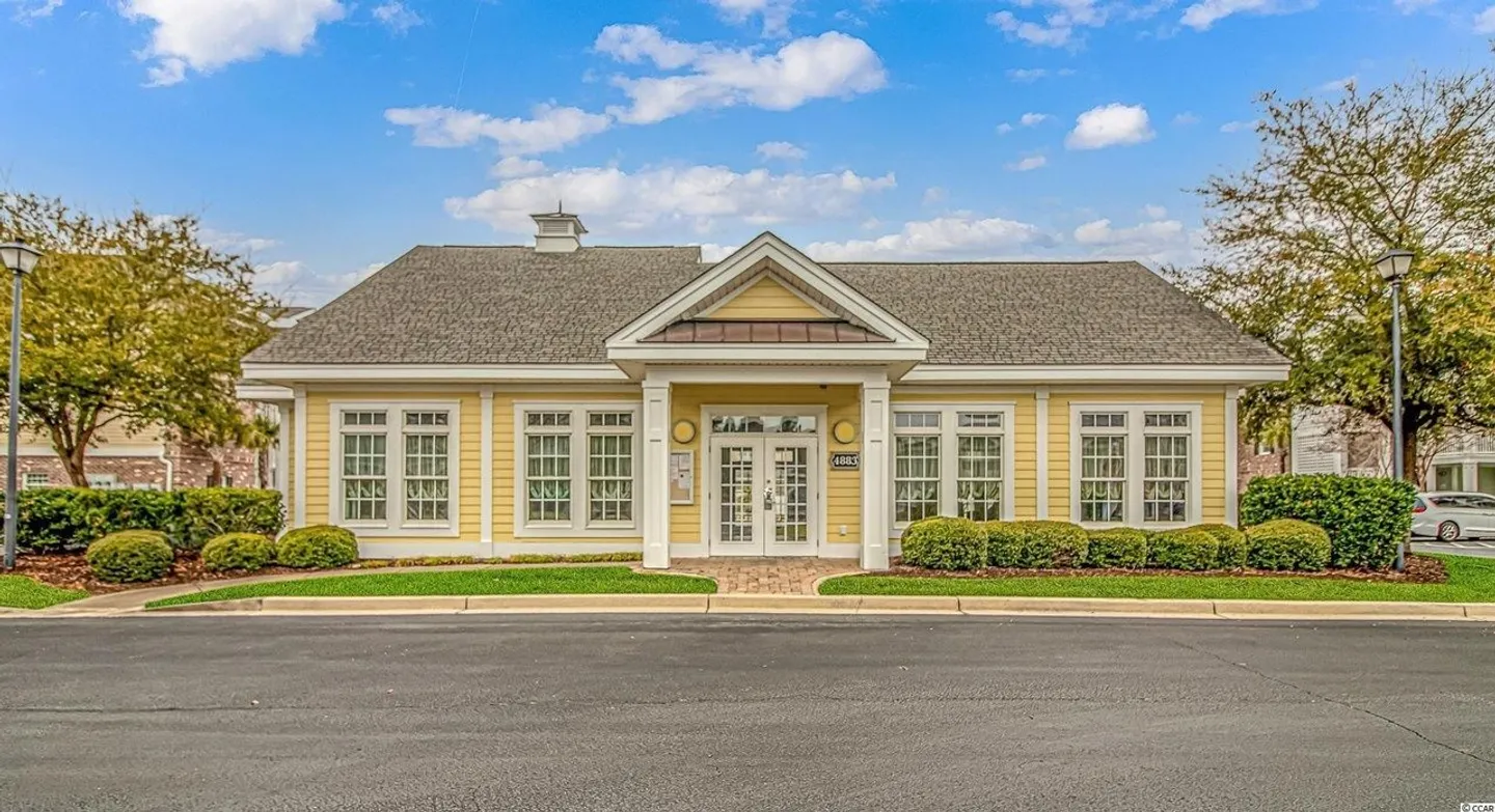 Front exterior view of a single-story yellow clubhouse building with white trim, a covered entrance, and a paved road in the foreground.