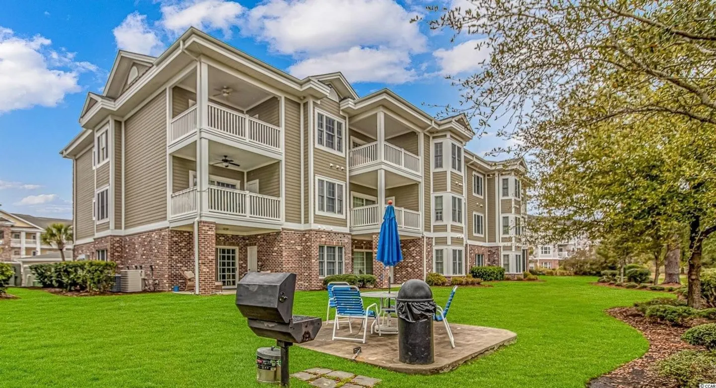 Exterior of a multi-story apartment building with balconies, green lawn, barbecue grill, and outdoor table.