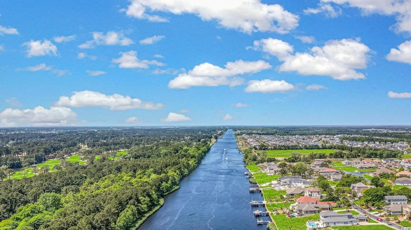 Aerial view of a long, straight canal bordered by dense green trees on one side and a residential neighborhood with boat docks on the other under a bright blue sky.
