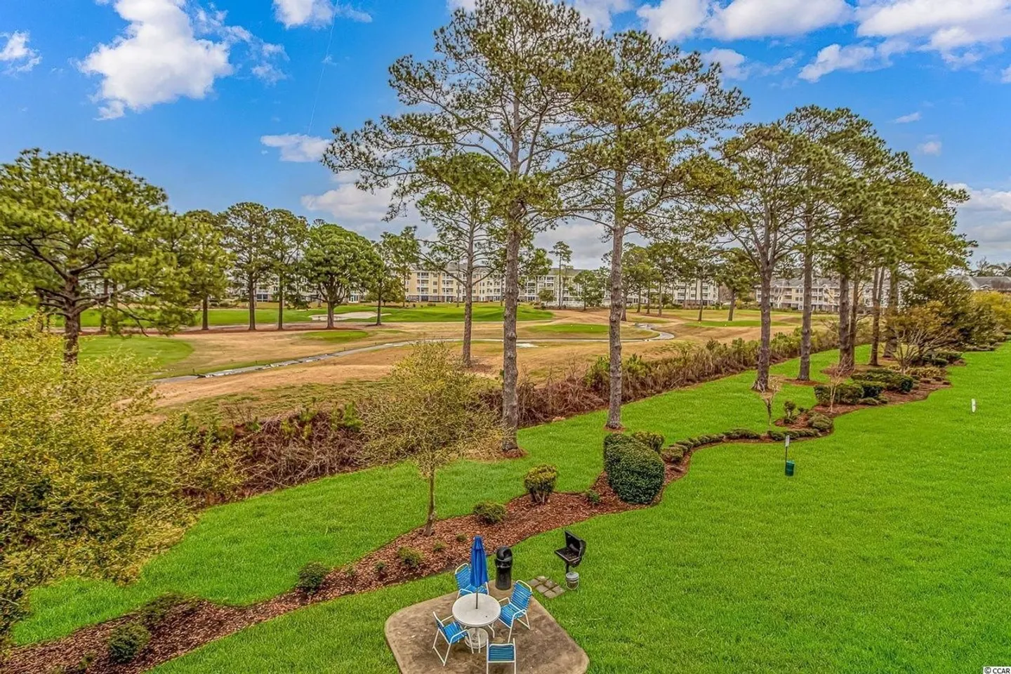 Elevated view of a lush green lawn, small patio with outdoor dining set, tall pine trees, and a golf course under a partly cloudy sky.