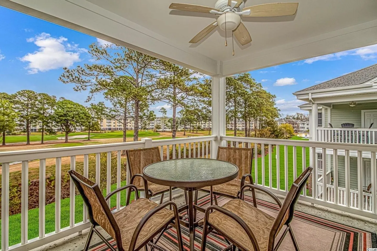 Covered balcony with a table, chairs, and ceiling fan overlooking green lawns and trees.