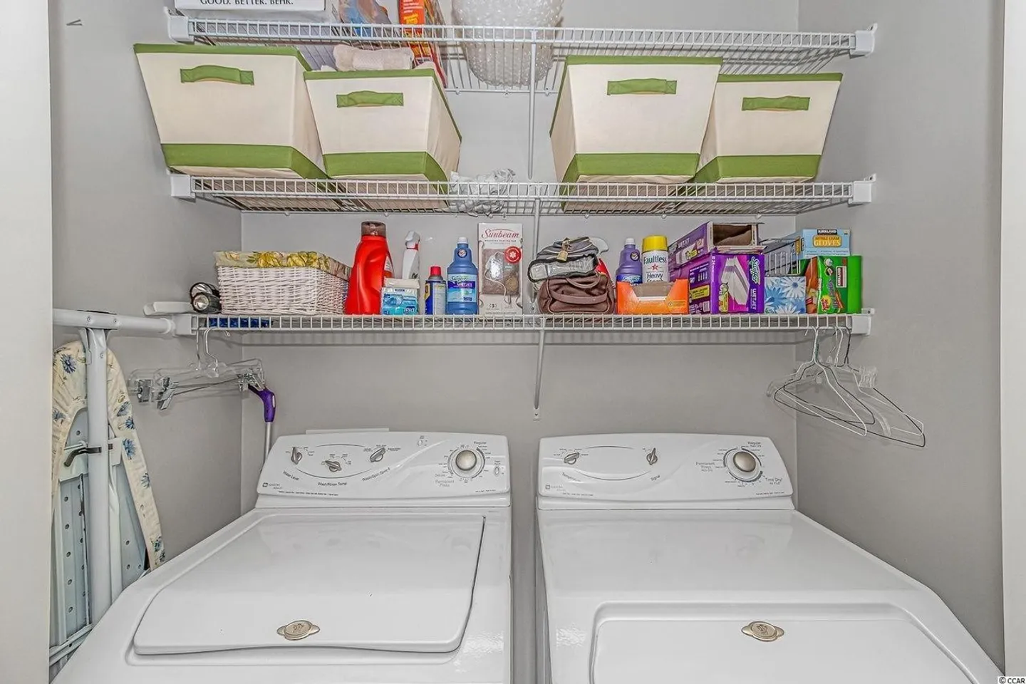 Laundry area featuring a side-by-side white washer and dryer beneath wire shelving with storage baskets and supplies.