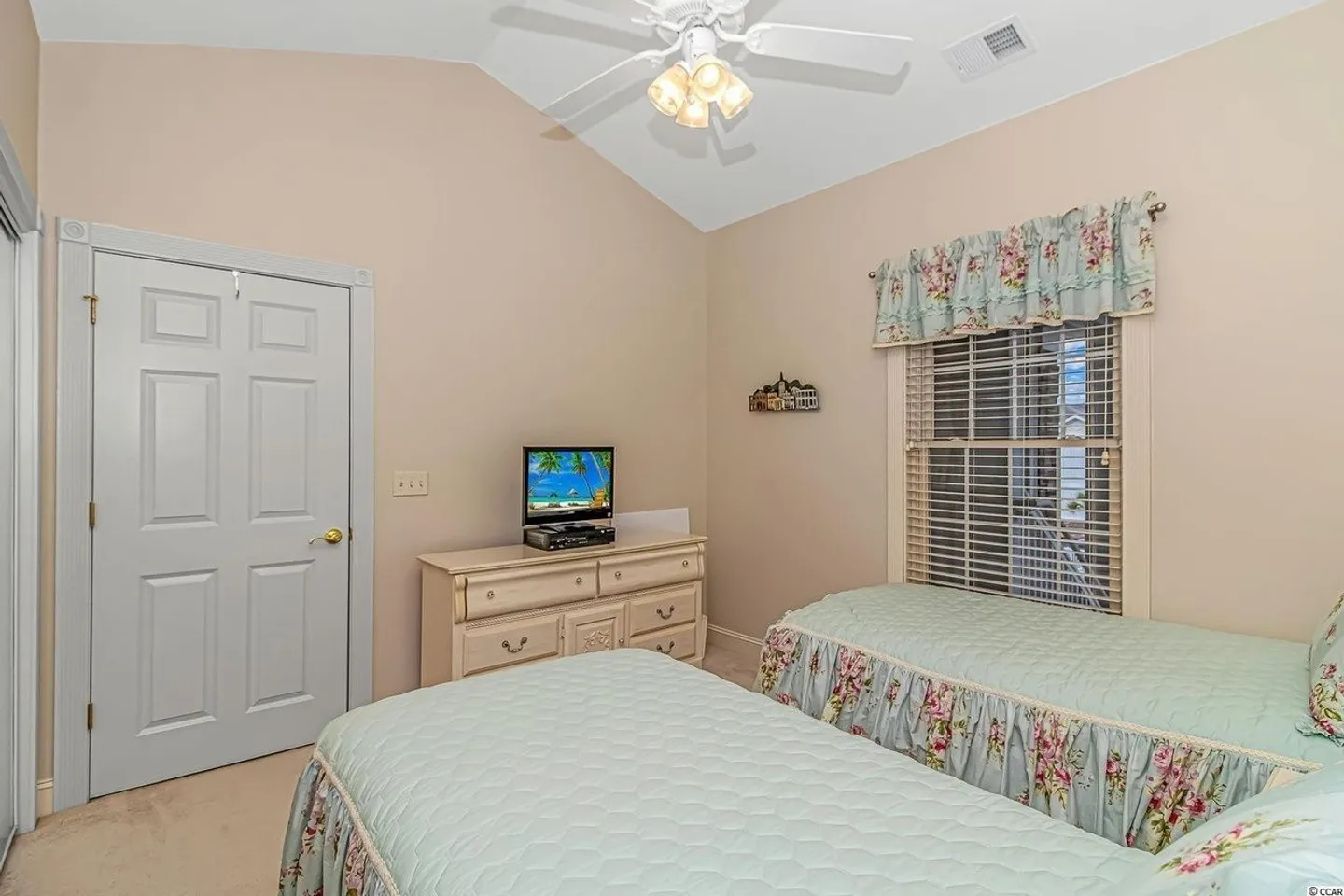 Bedroom with two twin beds featuring light blue and floral bedspreads, a dresser with a television, and a window with blinds.