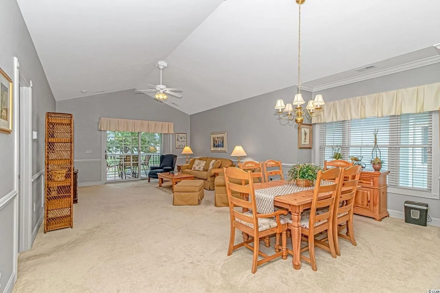Open living and dining area with carpeted floors, vaulted ceiling, wooden dining table, and living room furniture leading to a balcony.