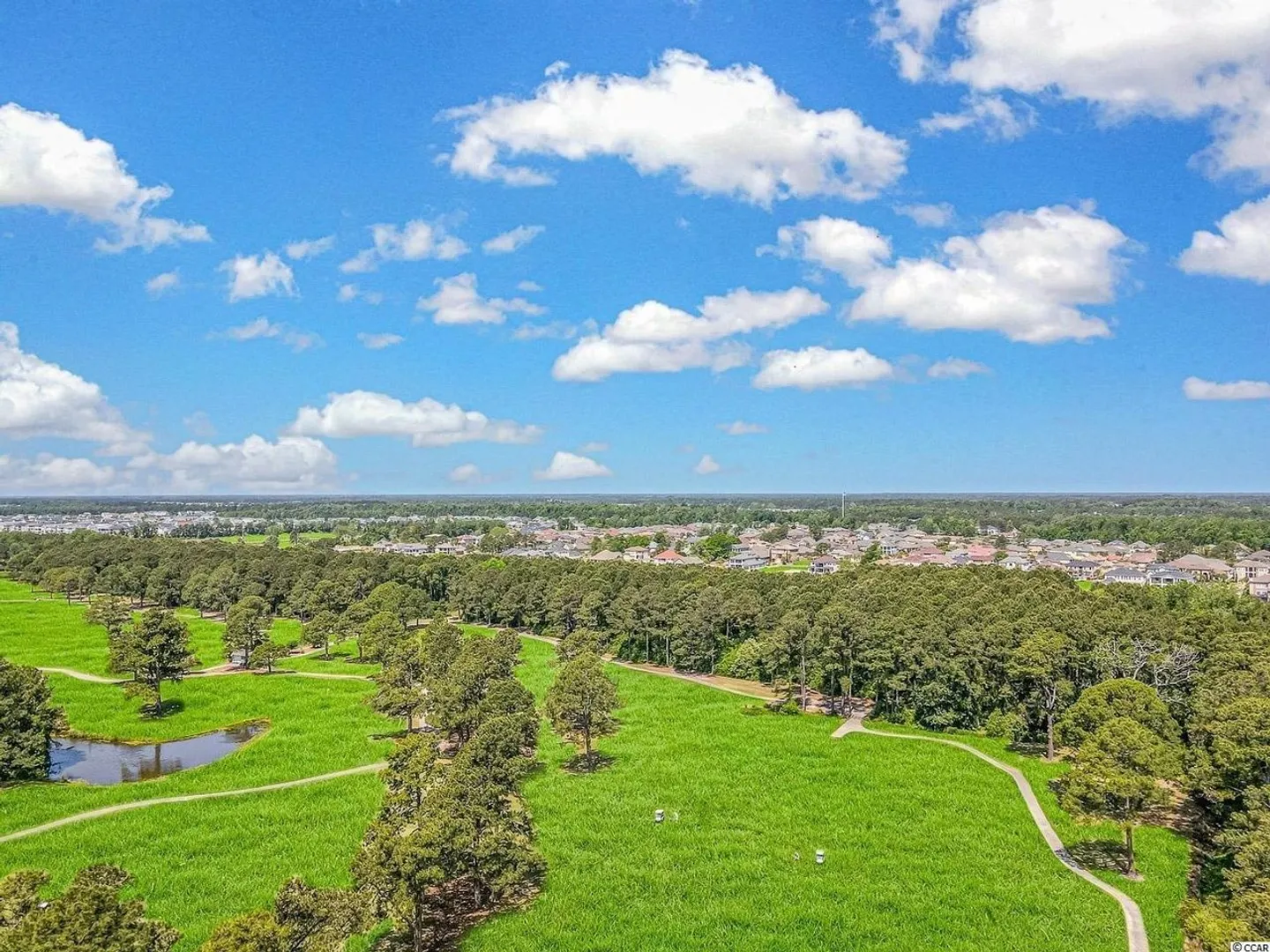 Aerial view of a green golf course with paved paths, trees, a pond, and a distant neighborhood under a blue, cloudy sky.