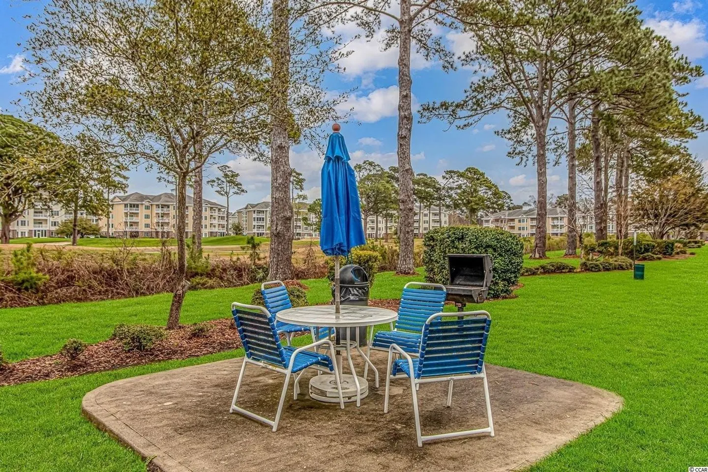 Outdoor picnic area with a concrete pad, patio table, blue chairs, folded umbrella, and standing charcoal grill surrounded by a grassy lawn and trees.