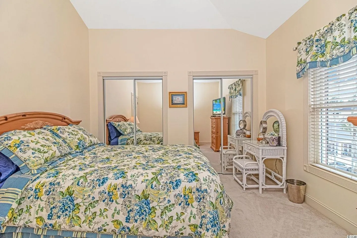 Bedroom with a bed in floral bedding, mirrored closet doors, a white wicker vanity table, and a window with blinds.
