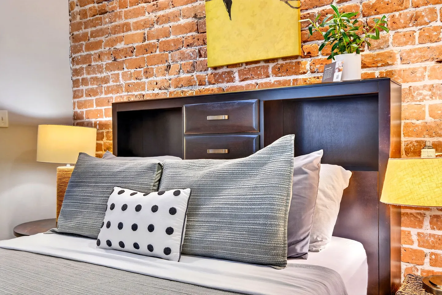Close-up of a bed set against an exposed brick wall with a dark wood bookcase headboard and lamps on either side.