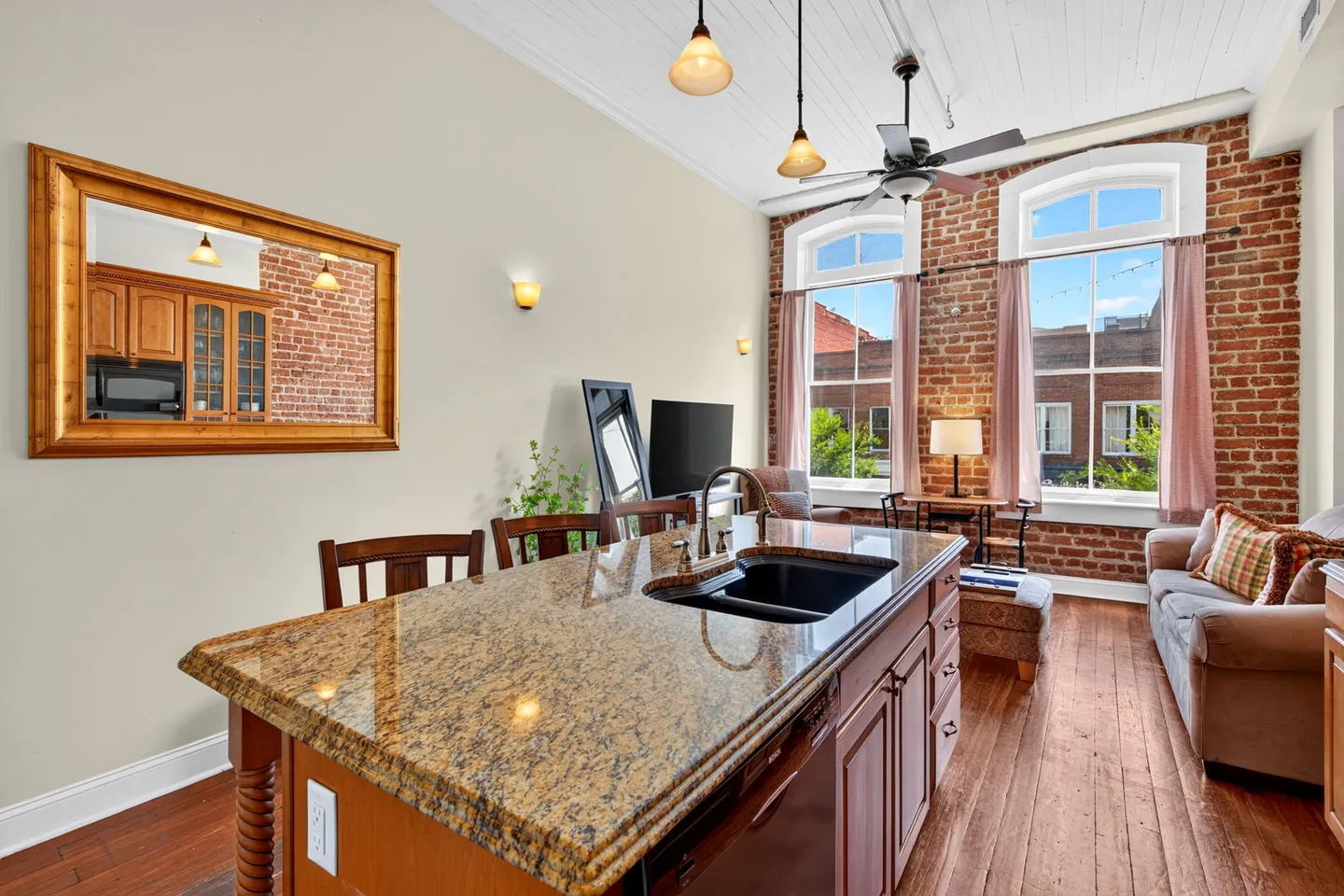 Open concept living area and kitchen with an island, hardwood floors, high ceilings, and exposed brick walls with tall arched windows.