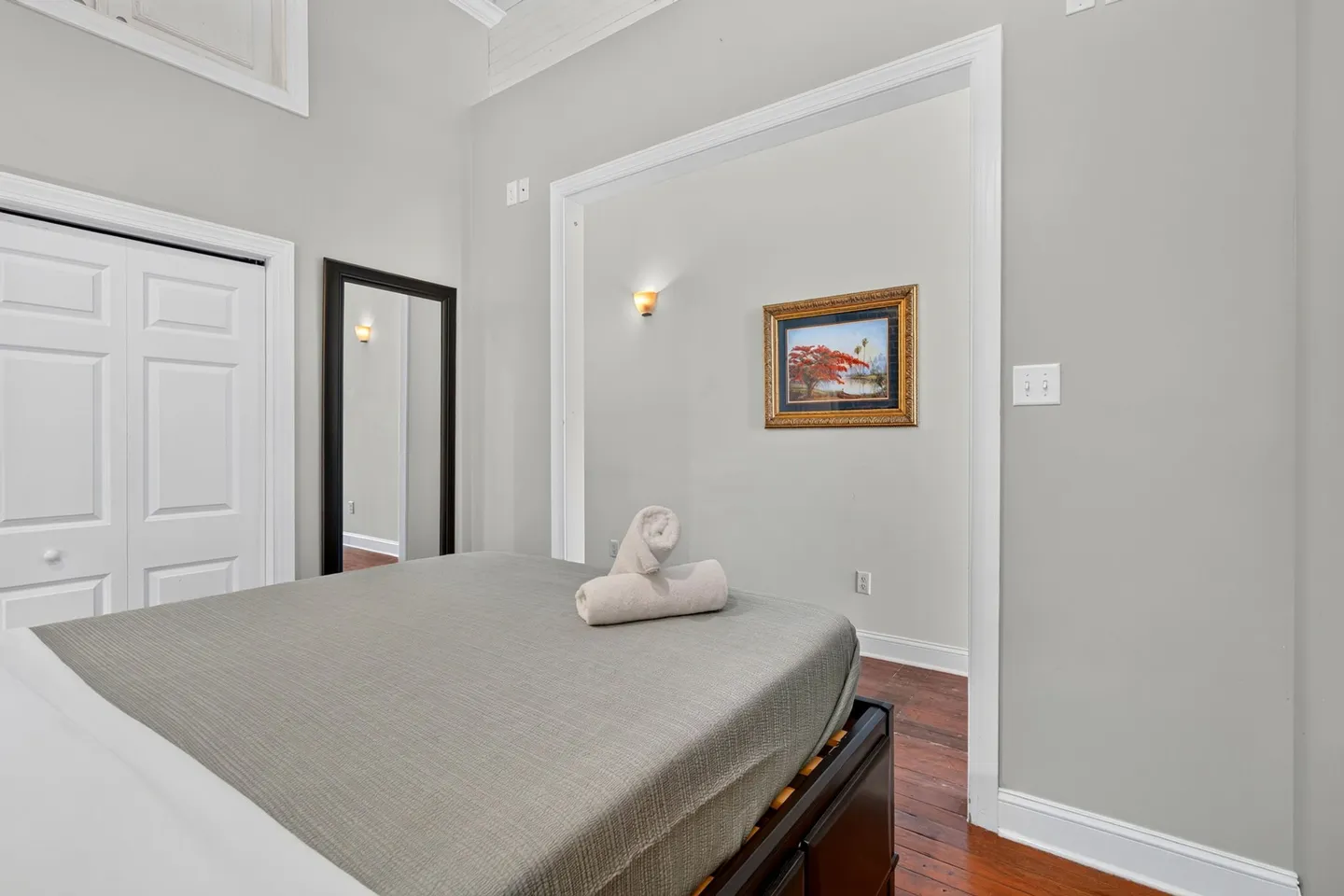 Bedroom with a made bed, wood floors, a full-length mirror, and framed wall art.
