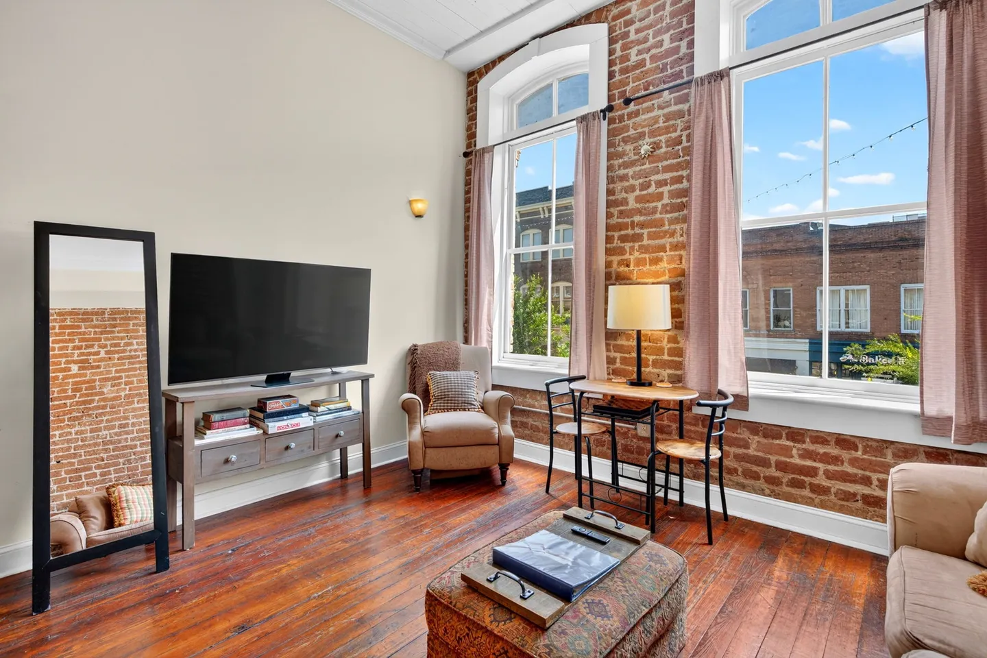 Living room with exposed red brick walls, high ceilings, large arched windows, wood floors, a flat-screen TV on a console, an armchair, and a small bistro table.