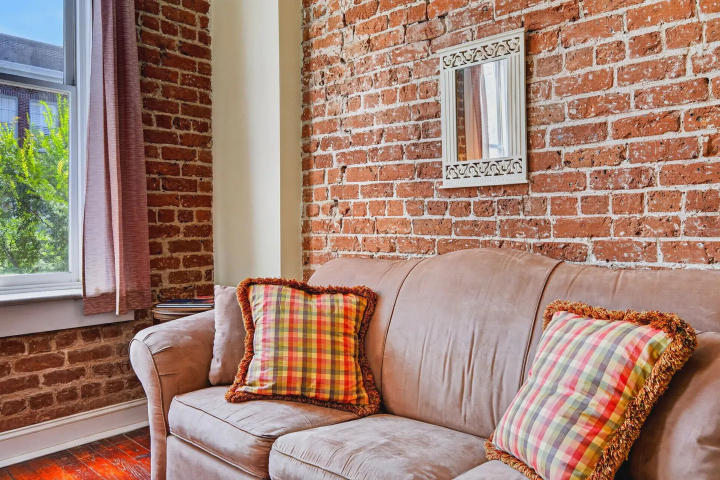 A tan sofa with plaid throw pillows set against an exposed red brick wall beside a window.