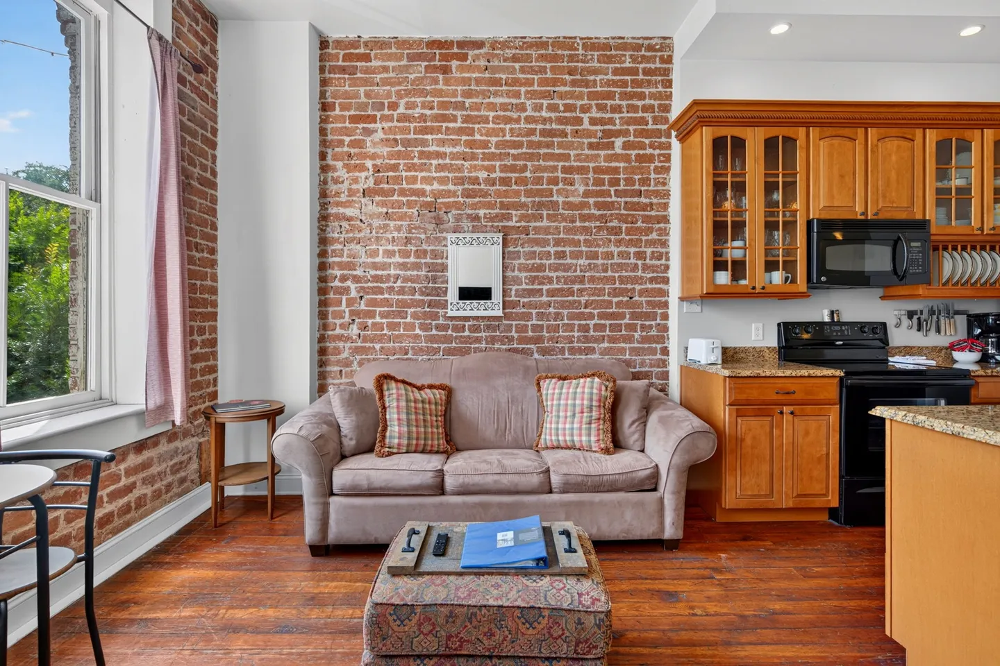 Living and kitchen area featuring an exposed red brick wall, a beige sofa with plaid pillows, an ottoman coffee table, and wood cabinets.