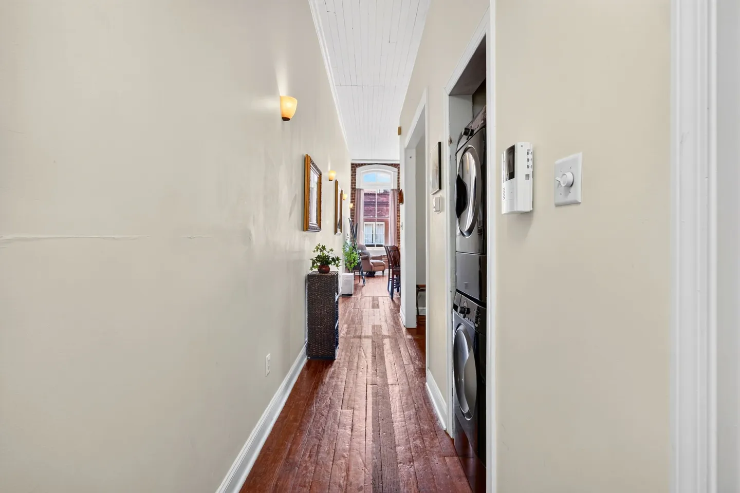 A long hallway with wood flooring, high ceilings, wall sconces, and a stacked washer and dryer in a recessed nook.
