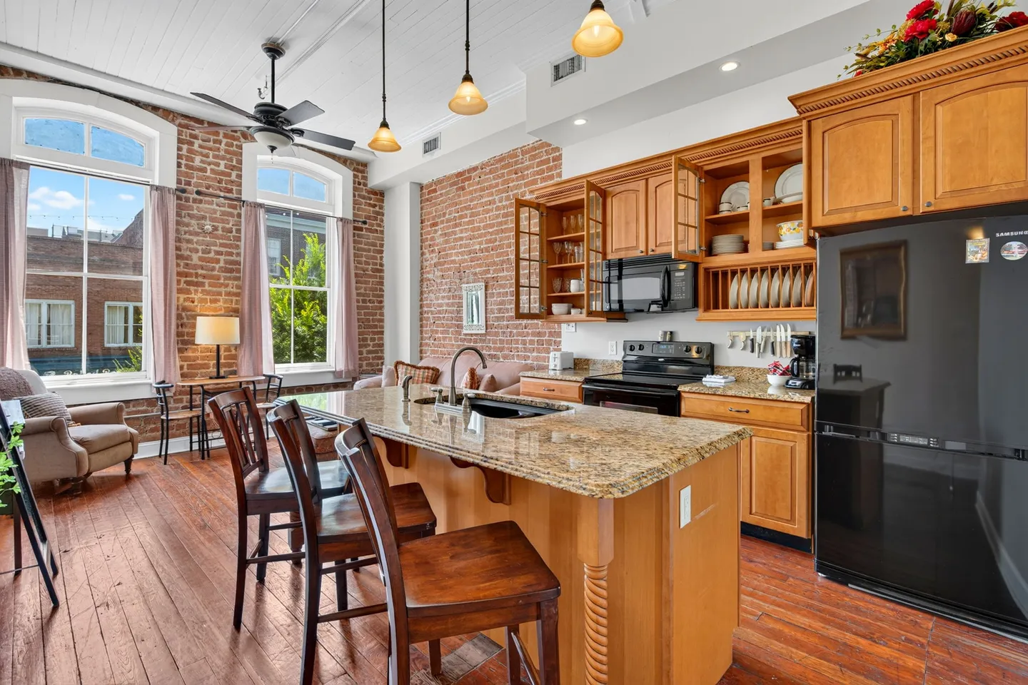 Open-concept kitchen and living area featuring exposed brick walls, hardwood floors, large arched windows, and an island with bar seating.