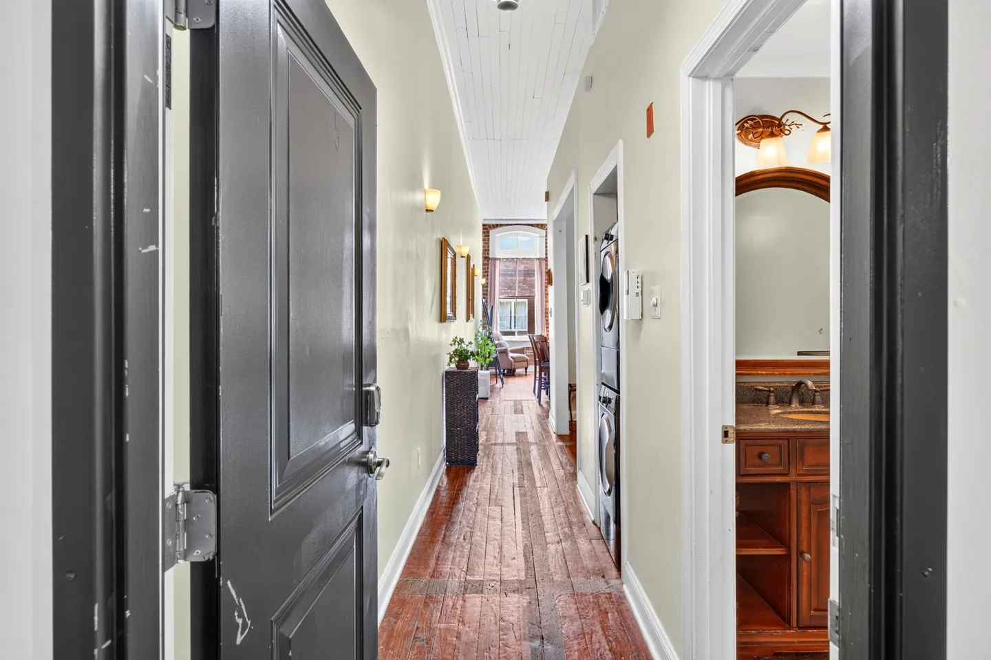 A long hallway with hardwood floors leading past an open bathroom and laundry nook toward a bright living area.