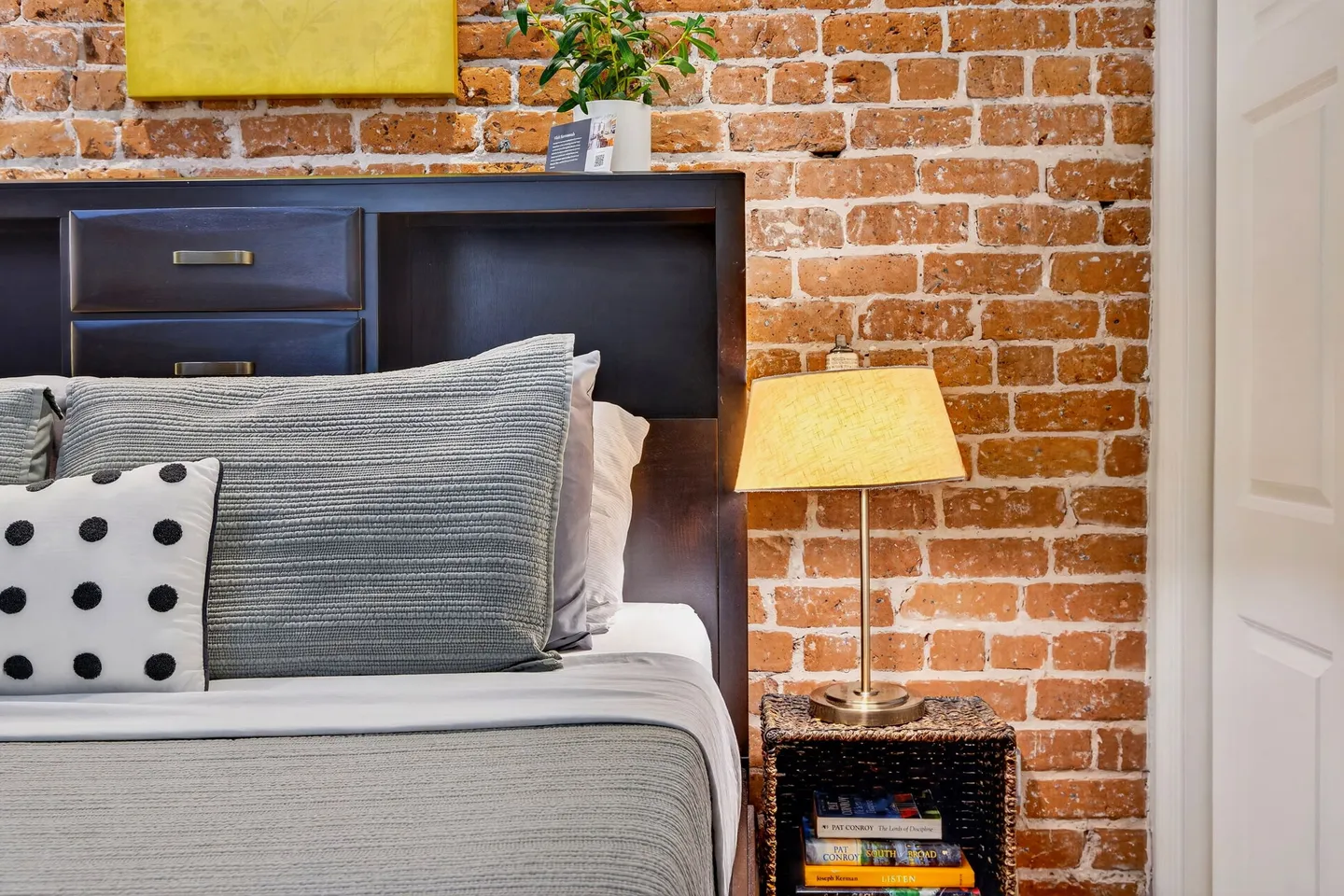 Bedroom detail featuring a dark wooden headboard with built-in cubbies against an exposed red brick wall, next to a small nightstand with a lamp.