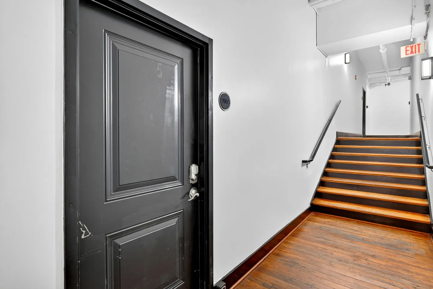 A black unit entry door in a hallway next to a flight of stairs with wood steps and a black handrail.
