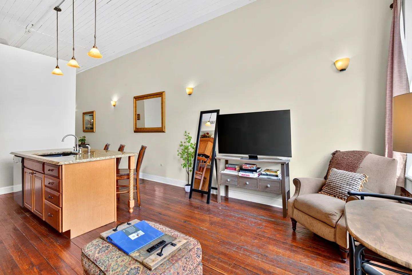Open living area and kitchen island featuring wood floors, high ceilings, a flat-screen TV on a media console, and an armchair.