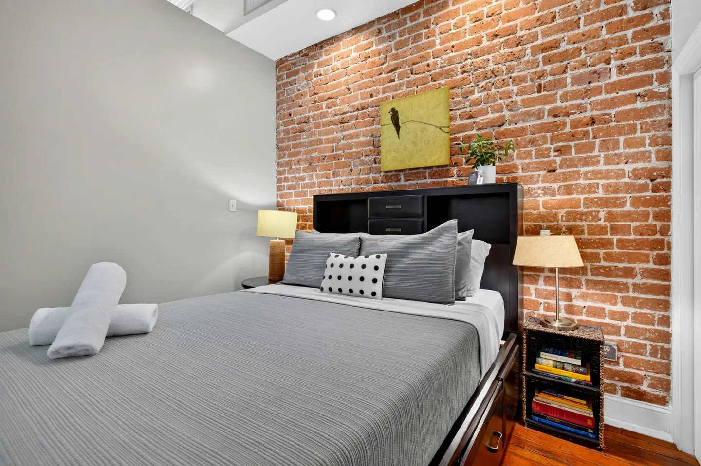 Bedroom featuring an exposed red brick accent wall behind a bed with grey bedding and two lit bedside lamps.