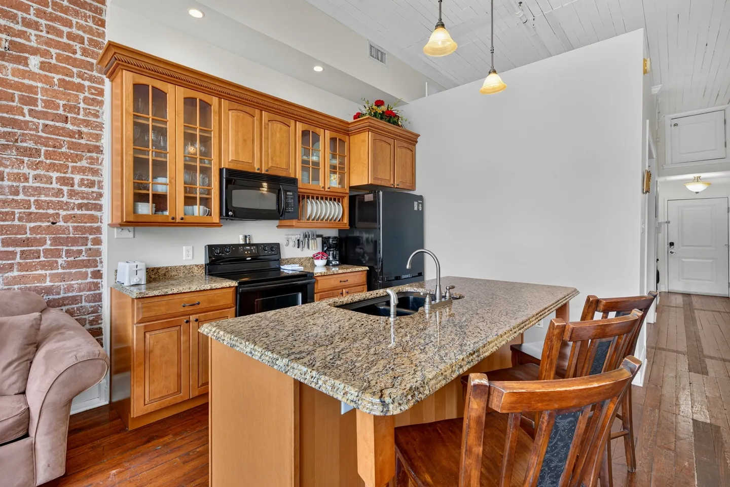 Open-concept kitchen with wooden cabinetry, granite countertops, an island with sink and barstools, and an exposed brick accent wall.