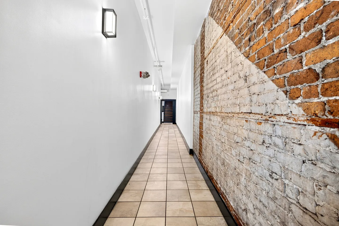 A long hallway with tiled flooring, a plain white wall on the left, and an exposed brick wall on the right.