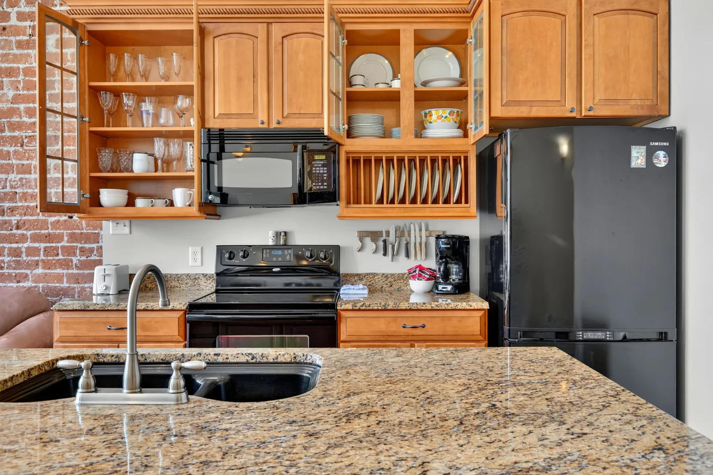 Kitchen featuring open wooden upper cabinets showing glassware and dishes, speckled granite countertops, and black appliances.