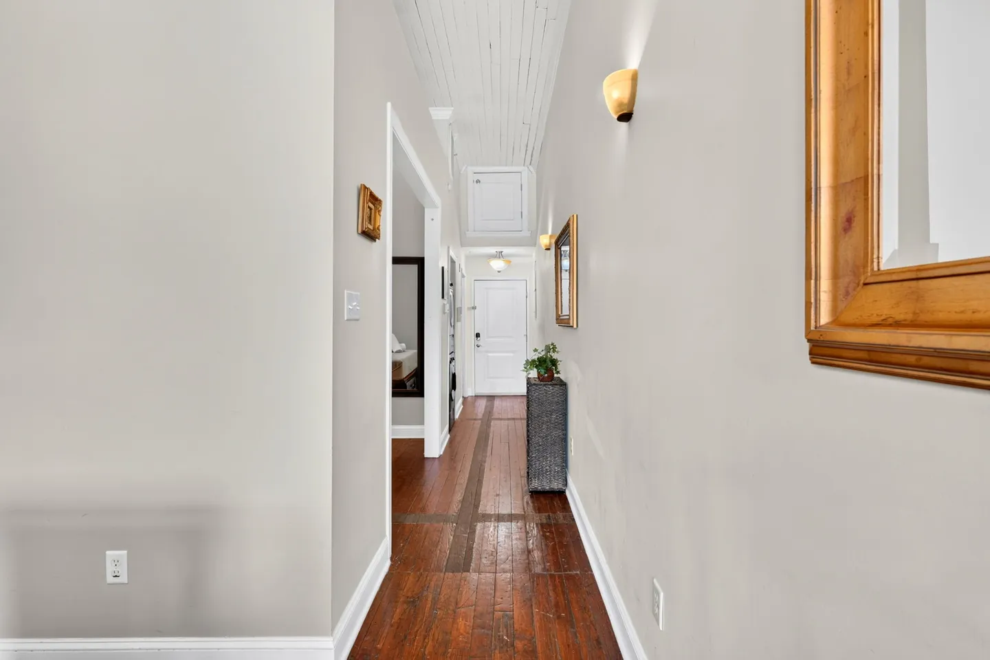 A long interior hallway featuring dark wood flooring, light-colored walls, a high ceiling, and a front entry door at the end.