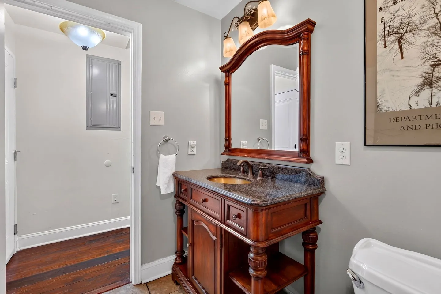 Bathroom vanity featuring a carved wood cabinet, dark countertop, framed mirror, and an overhead three-light fixture beside a doorway.