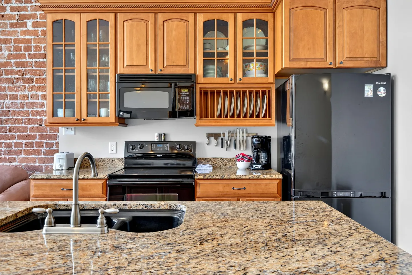 Kitchen featuring wood cabinetry, granite countertops, a black refrigerator, microwave, and stove, alongside an exposed brick wall.