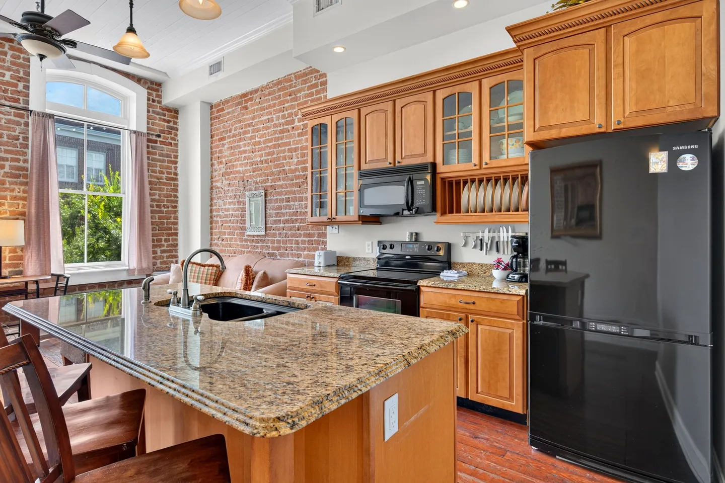 Kitchen with an exposed brick wall, granite-topped island, wooden cabinetry, and black appliances next to a living area.