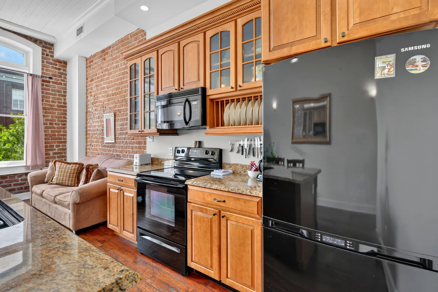 Kitchen with wooden cabinetry, black appliances, and granite-look counters opening to a living area with an exposed brick wall.