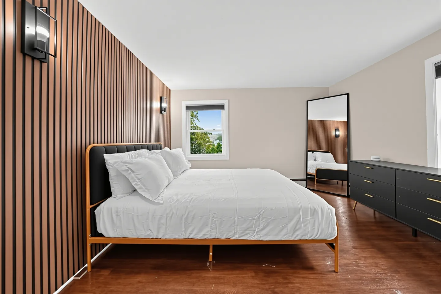 Bedroom featuring a bed with white linens, a vertical wood slat accent wall with sconces, and a large leaning mirror.