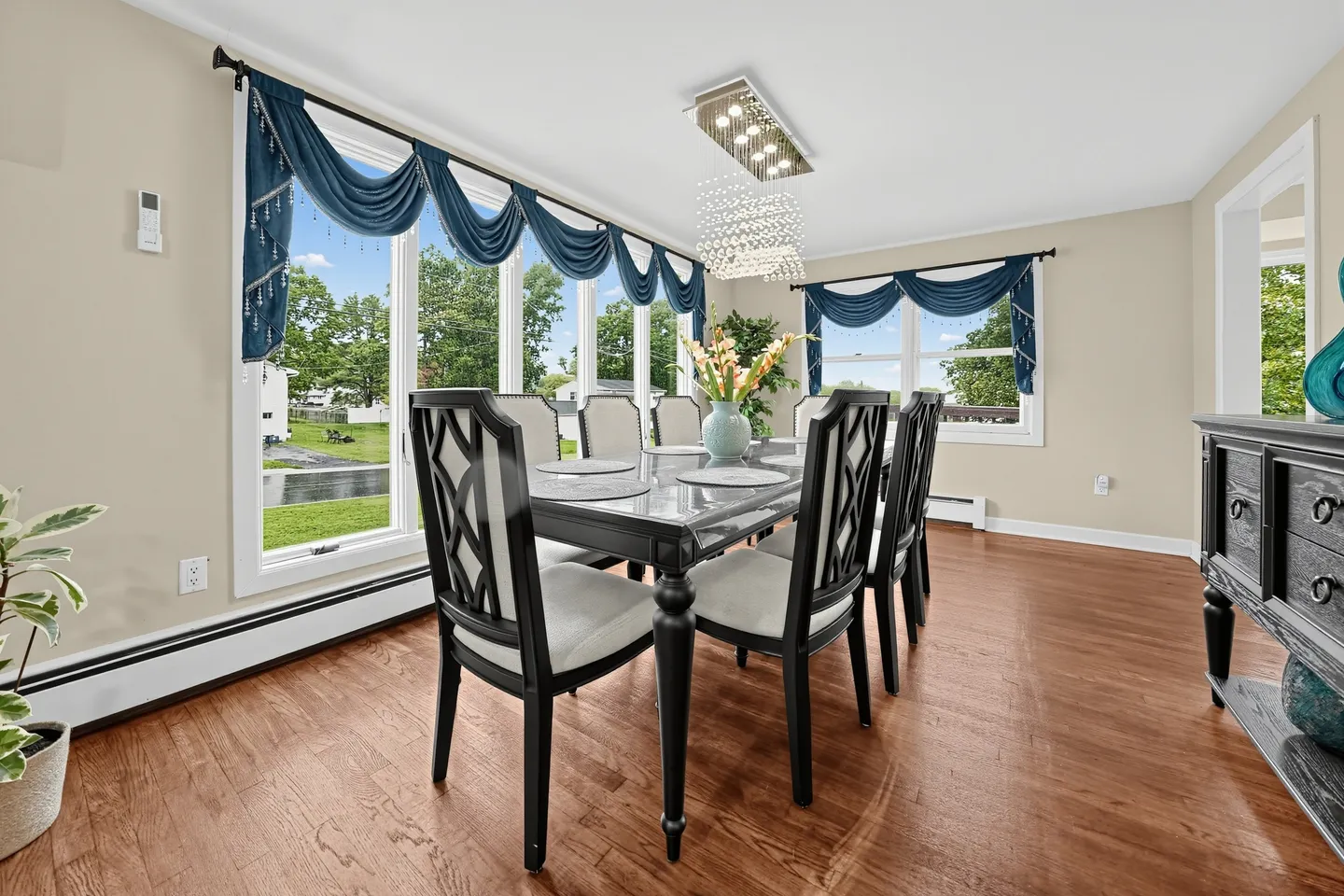 Dining room with a polished table, upholstered dark wood chairs, a crystal chandelier, and large windows with blue swag valances.