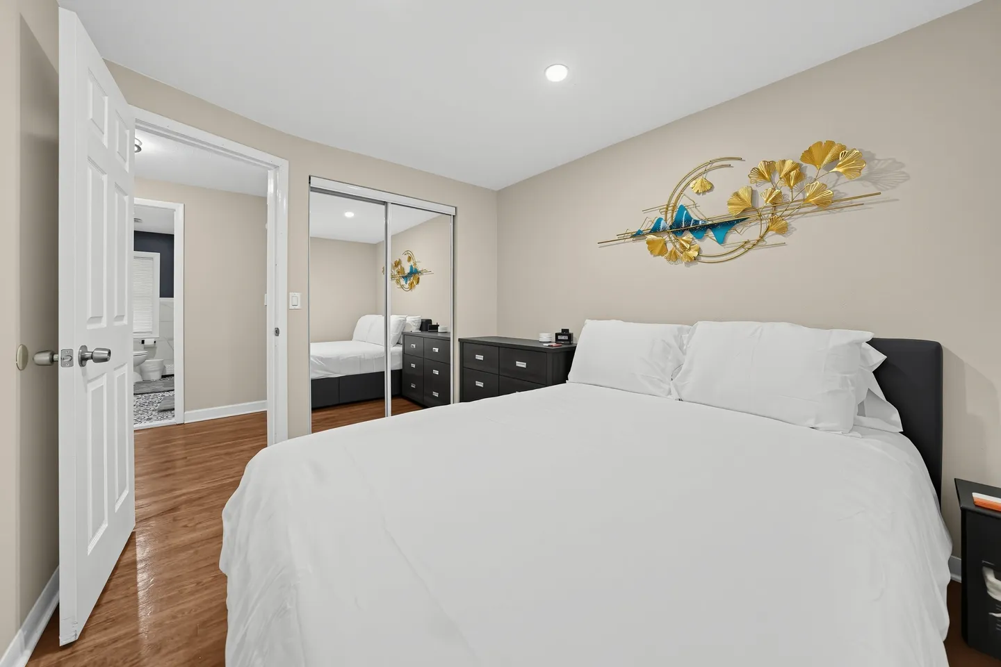 Bedroom with a bed made with white linens, mirrored closet doors, a dark dresser, and decorative wall art.