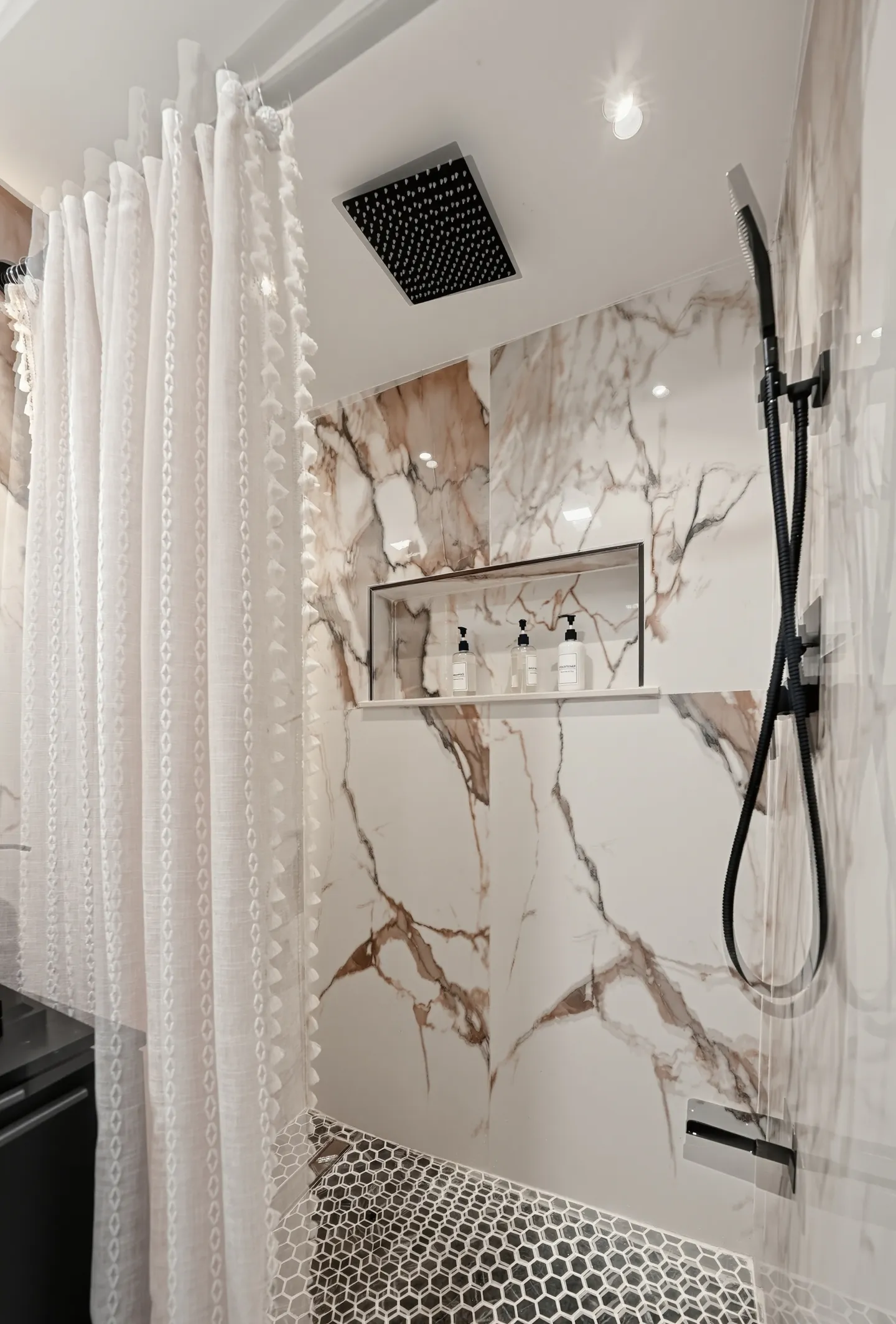 Walk-in shower with marble-look wall tiles, black hexagon floor tile, overhead rain showerhead, and a white curtain.