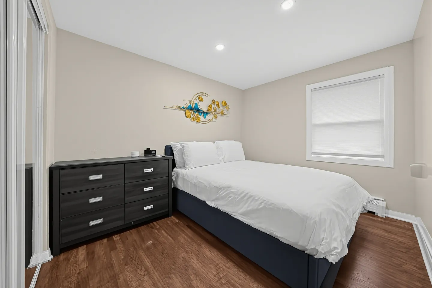 Bedroom with a bed made in white linens, a dark dresser, mirrored closet doors, and recessed ceiling lights.