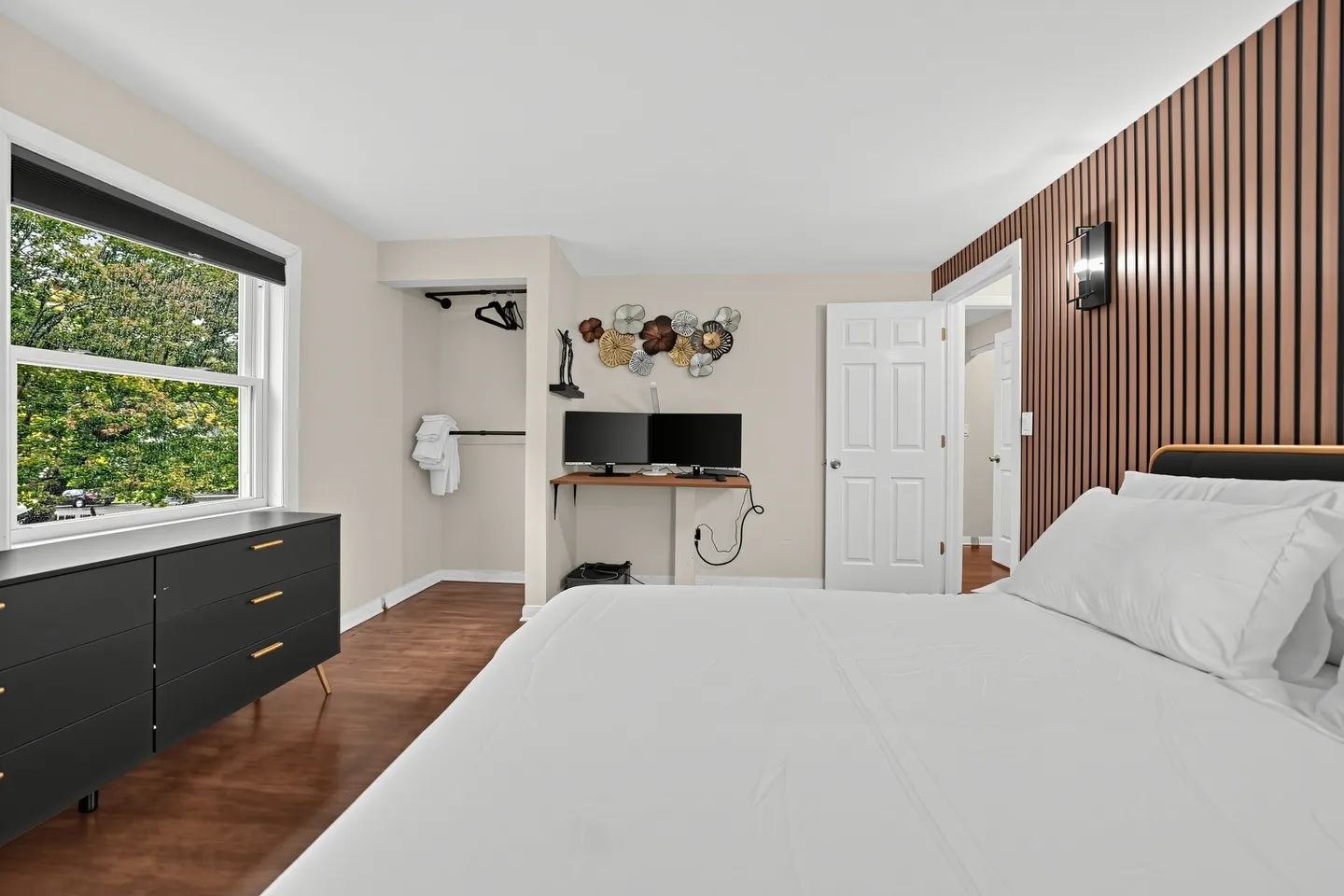 Bedroom with a white bed, wood slat accent wall, dark dresser, window, open closet space, and a small desk with dual monitors.