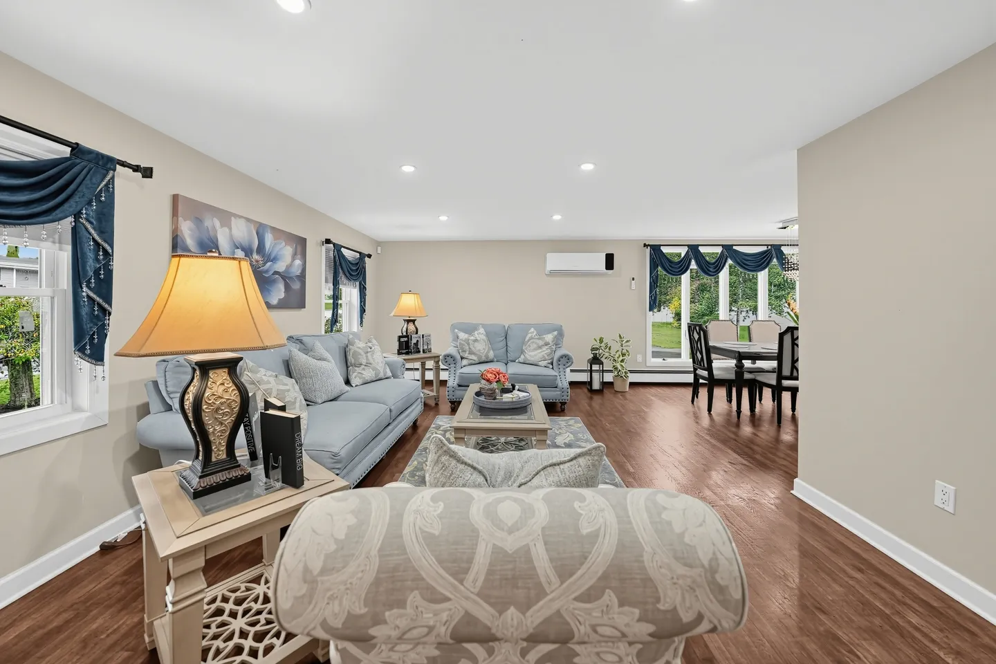 Open-concept living room with light blue sofas, an ornate table lamp, wood-look flooring, and an adjoining dining area.