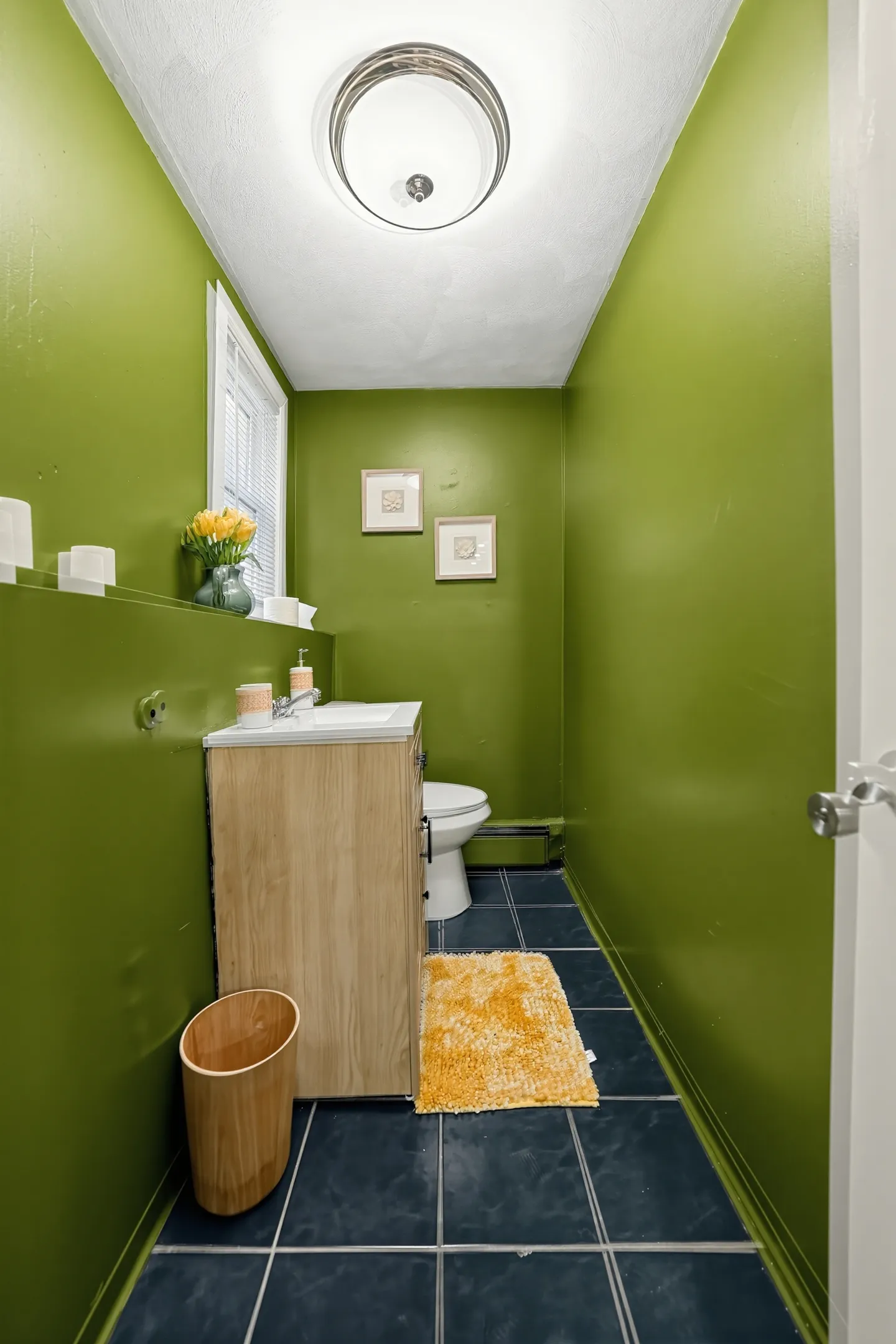 A long, narrow bathroom with bright green walls, dark tiled floors, a light wood vanity, and a toilet in the back.