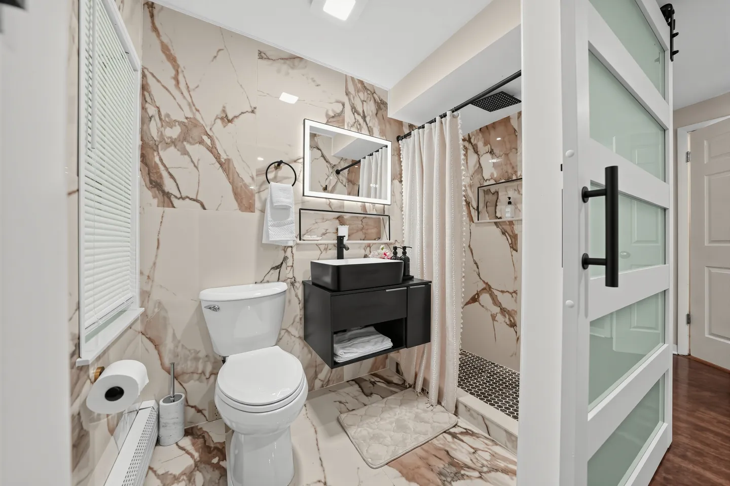Bathroom with marble-look wall tiles, a toilet, a black vessel sink vanity with an illuminated mirror, and a shower with a curtain.