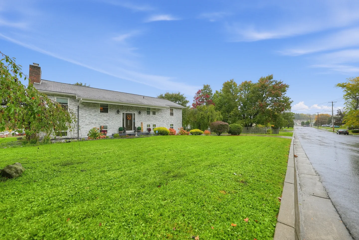 Exterior view of a split-level home with a large green lawn beside a wet asphalt street under a blue sky.