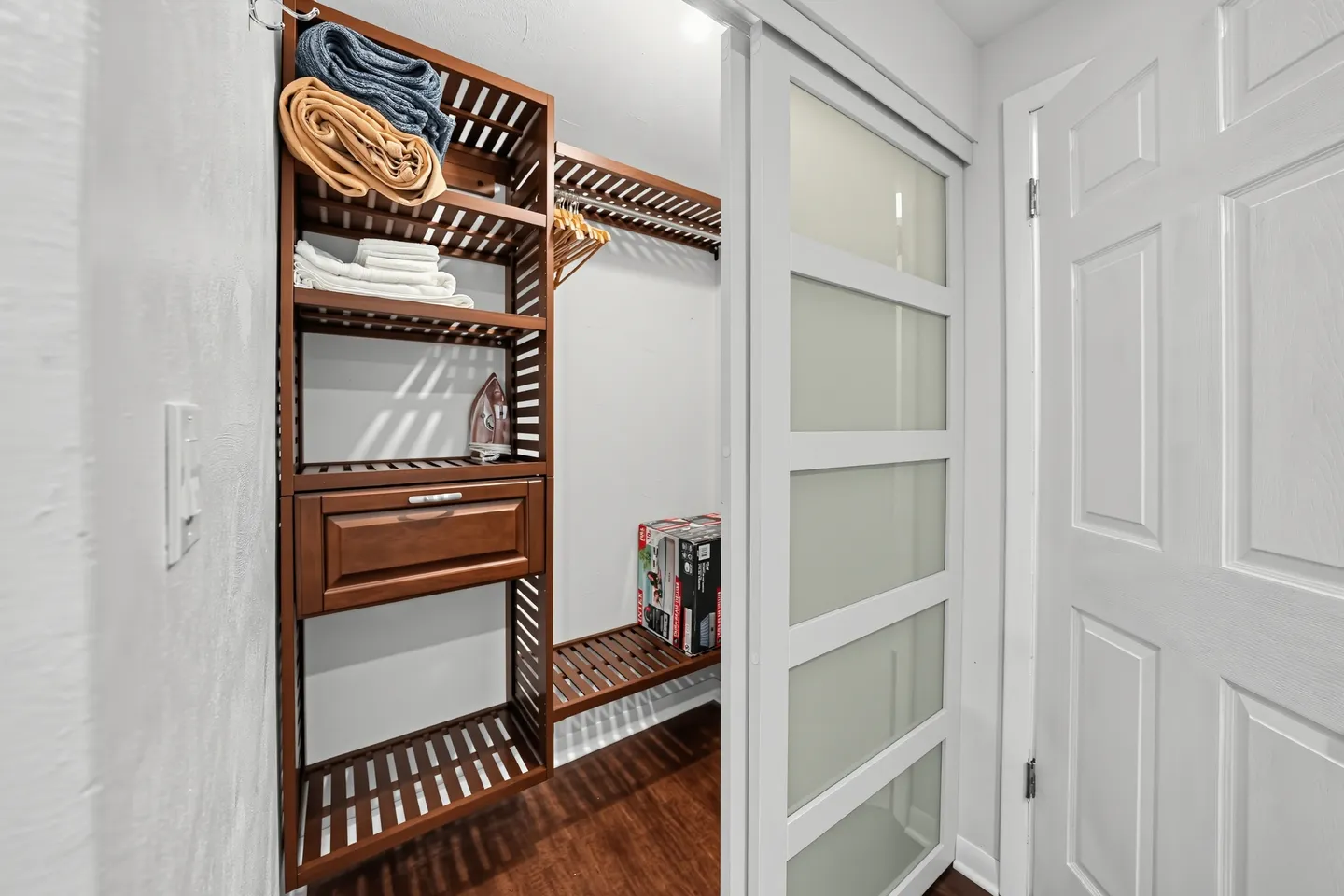 A walk-in closet behind a frosted glass sliding door, featuring built-in wooden shelving, a drawer, and stored linens.