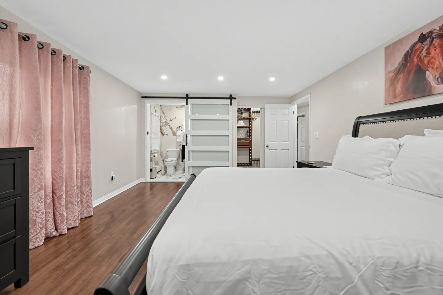 Bedroom with a white-linened bed, wood-style flooring, a sliding barn-style door, and views into an en-suite bathroom and closet.