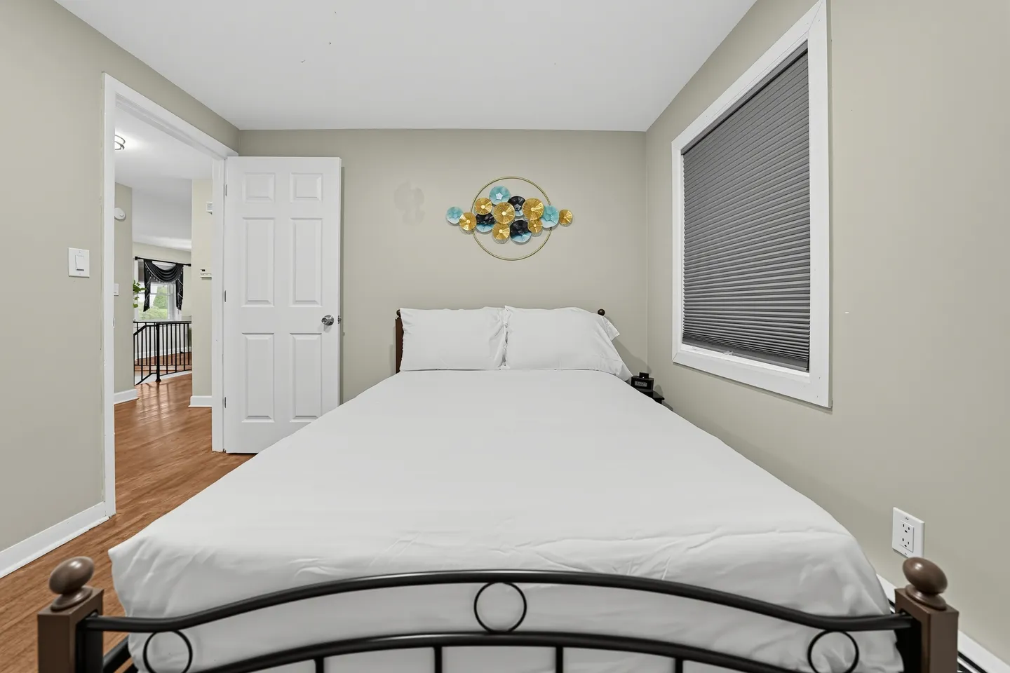 Bedroom with a bed made with white linens, metal headboard, a window with closed blinds, and an open door showing a hallway.