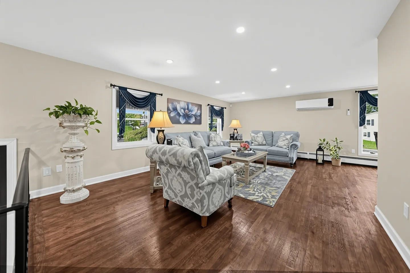 Spacious living room with hardwood floors, light blue sofas, an accent armchair, and multiple windows with dark drapery.