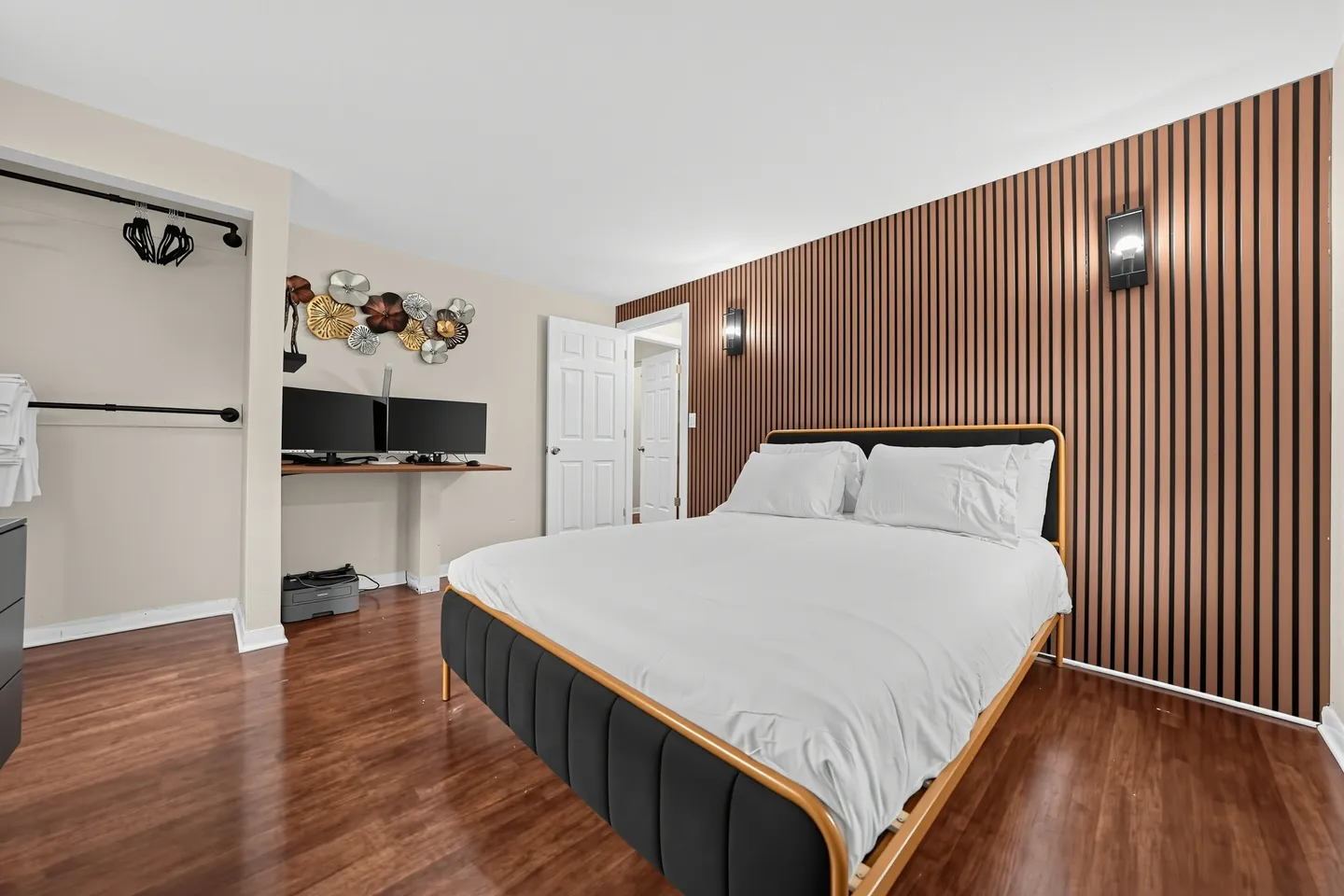 Bedroom with wood-look flooring, a bed with a fluted black and wood frame, a vertical wood slat accent wall with sconces, and a desk workspace.